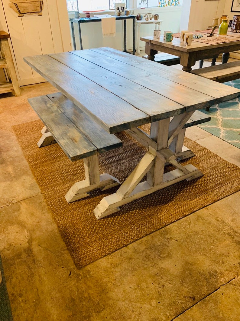 May include: A rustic farmhouse-style dining table and bench set. The table features a distressed gray-blue tabletop and white-painted trestle-style legs. The bench mirrors the table's design. The set is placed on a woven brown rug.