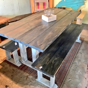 May include: A large, rustic wooden dining table with two matching benches. The table and benches are made of gray-stained wood and have white-painted legs. A small wooden box with glasses sits on the table. The table is set on a patterned rug.