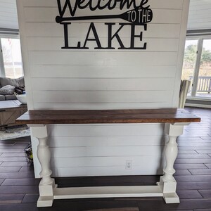 May include: A console table featuring a rectangular dark brown wooden top supported by two white painted pedestal legs with a matching white base. The wall behind the table is white shiplap with black text reading Welcome to the Lake.
