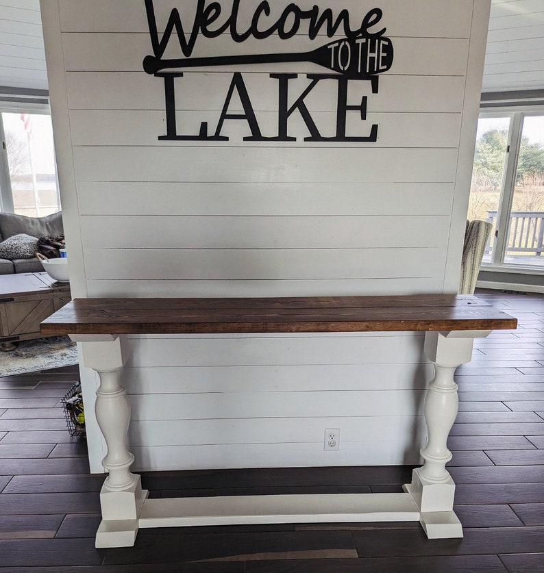 May include: A console table featuring a rectangular dark brown wooden top supported by two white painted pedestal legs with a matching white base. The wall behind the table is white shiplap with black text reading Welcome to the Lake.