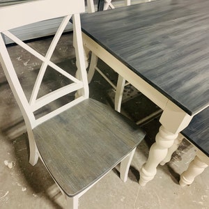 K&ouml;nnte beinhalten: Ein Esszimmerset bestehend aus einem rechteckigen Tisch und St&uuml;hlen mit wei&szlig;en Rahmen und dunkelgrau gebeizten Holzoberfl&auml;chen. Die St&uuml;hle haben ein X-f&ouml;rmiges R&uuml;ckendesign und die Tisch- sowie Stuhlbeine weisen gedrechselte Details auf.