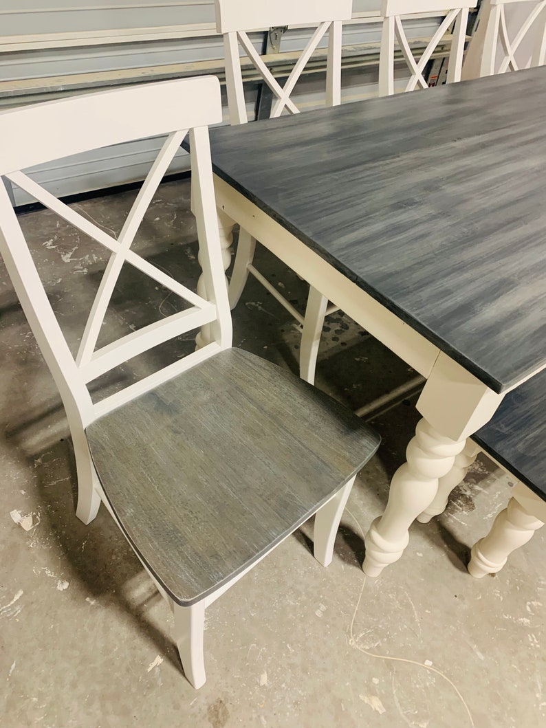 K&ouml;nnte beinhalten: Ein Esszimmerset bestehend aus einem rechteckigen Tisch und St&uuml;hlen mit wei&szlig;en Rahmen und dunkelgrau gebeizten Holzoberfl&auml;chen. Die St&uuml;hle haben ein X-f&ouml;rmiges R&uuml;ckendesign und die Tisch- sowie Stuhlbeine weisen gedrechselte Details auf.