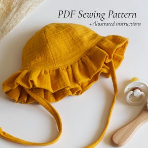 May include: A mustard yellow sun hat with a ruffled brim and long fabric ties. The text PDF Sewing Pattern + illustrated instructions is visible above the hat. A white pacifier and a wooden handle are positioned to the right of the hat.