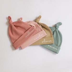 Peut inclure: Trois bonnets de b&eacute;b&eacute; personnalis&eacute;s en rose, beige et vert avec les noms "Willow", "Amelia" et "Oscar" brod&eacute;s dessus.
