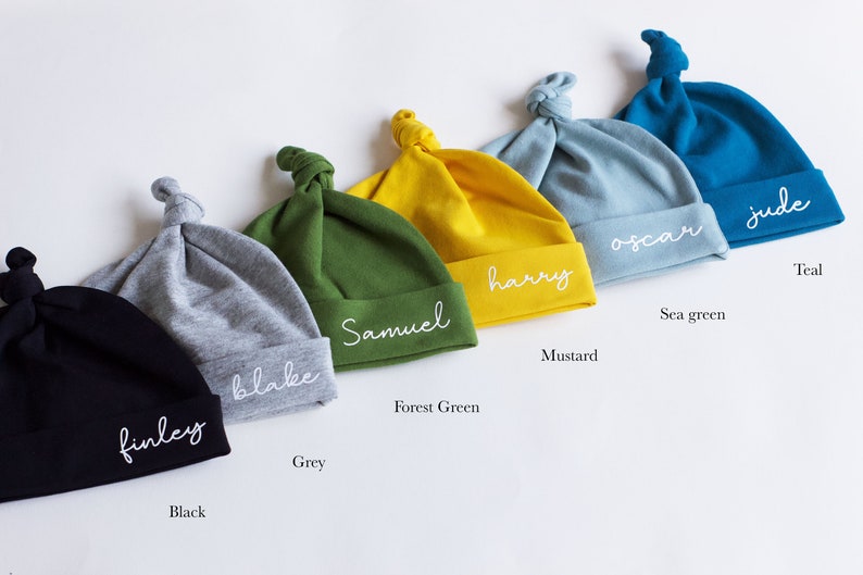 May include: Six personalized baby hats in different colors, including black, grey, forest green, mustard, sea green, and teal. Each hat has a different name embroidered on it, including Finley, Blake, Samuel, Harry, Oscar, and Jude.