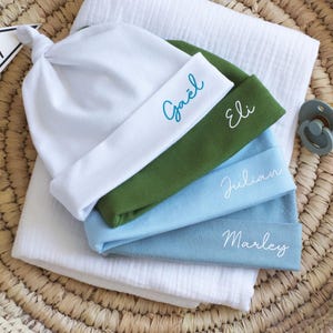 May include: Four baby hats stacked on top of each other. The hats are white, green, light blue, and blue. Each hat has a name embroidered on it: Ga&euml;l, Eli, Julian, and Marley.