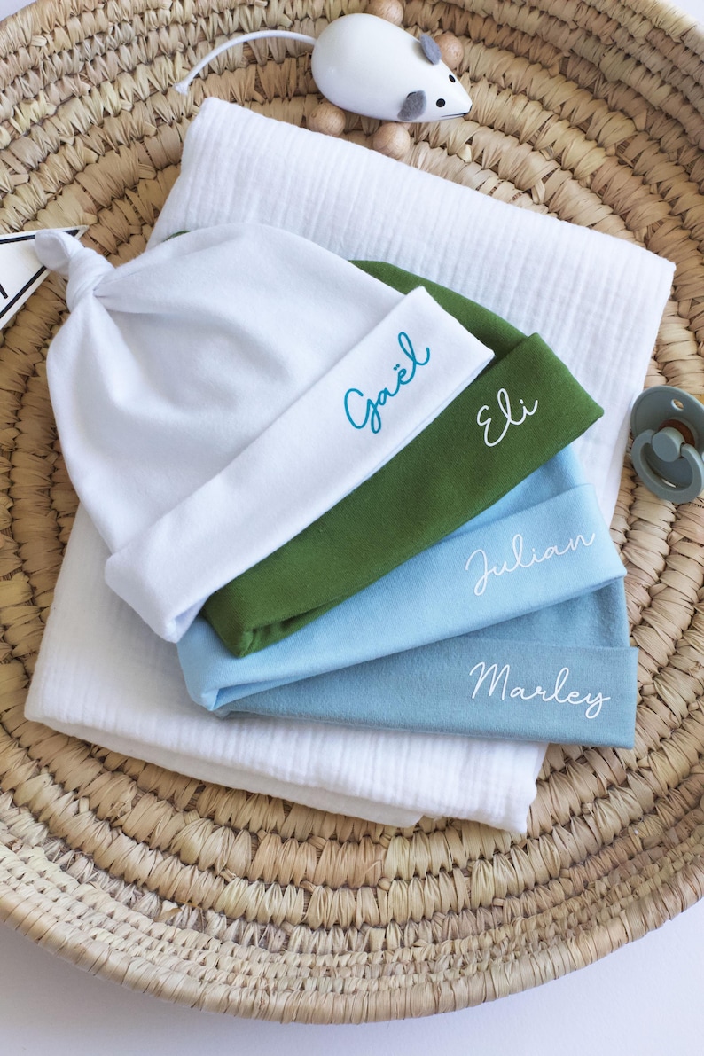 May include: Four baby hats stacked on top of each other. The hats are white, green, light blue, and blue. Each hat has a name embroidered on it: Ga&euml;l, Eli, Julian, and Marley.