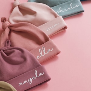 Peut inclure: Cinq bonnets pour b&eacute;b&eacute;s de couleurs diff&eacute;rentes avec des n&oelig;uds sur le dessus. Chaque bonnet a un nom diff&eacute;rent brod&eacute; en blanc sur le bord : Mia, Ella, Angela, Charlie et Jan.
