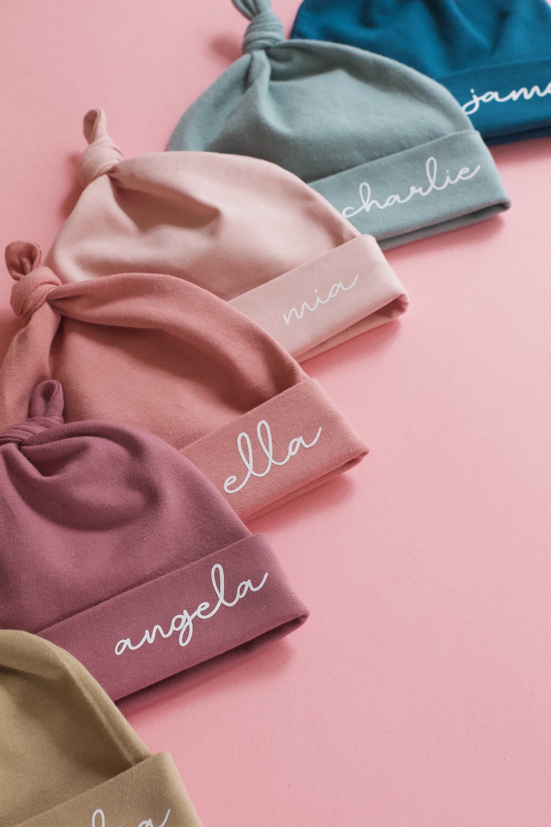 Peut inclure: Cinq bonnets pour b&eacute;b&eacute;s de couleurs diff&eacute;rentes avec des n&oelig;uds sur le dessus. Chaque bonnet a un nom diff&eacute;rent brod&eacute; en blanc sur le bord : Mia, Ella, Angela, Charlie et Jan.