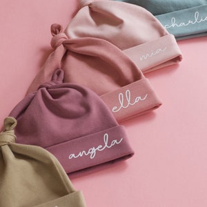 May include: Five different colored baby hats with knotted tops. Each hat has a different name embroidered on the bottom edge in white: Melia, Angela, Ella, Mia, and Charlie.