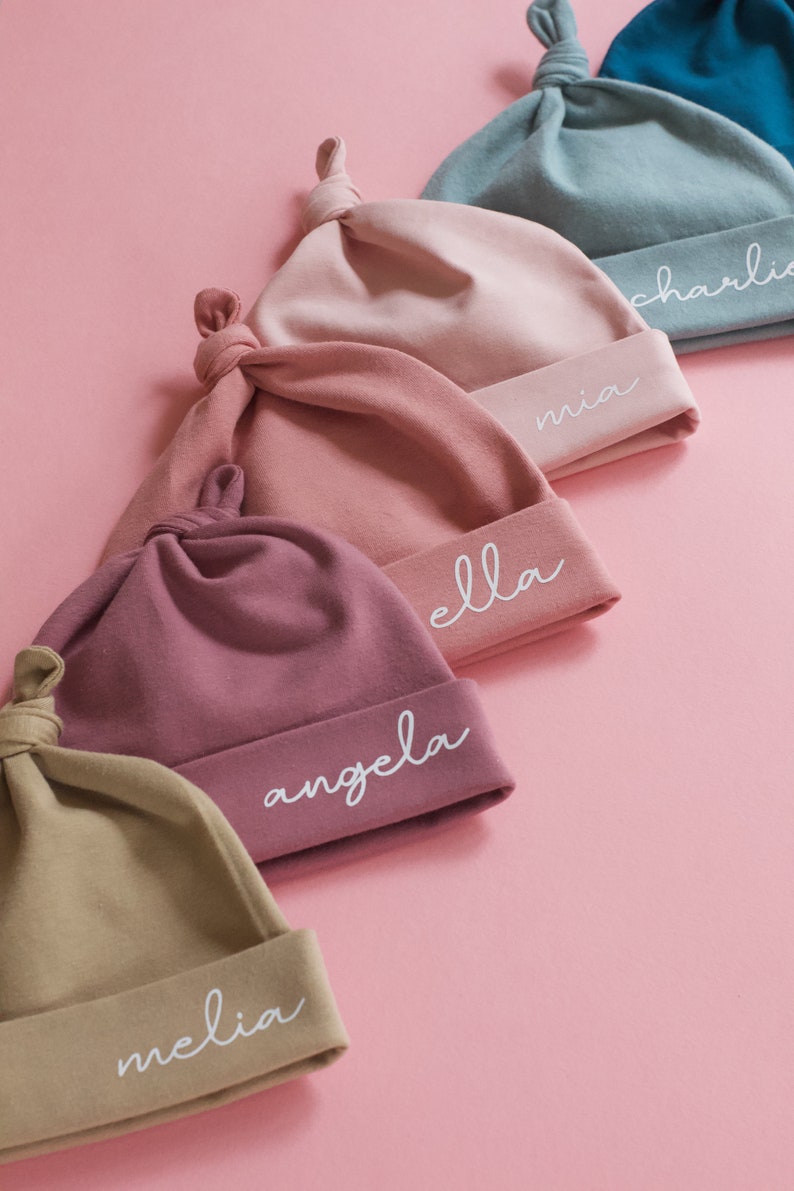 May include: Five different colored baby hats with knotted tops. Each hat has a different name embroidered on the bottom edge in white: Melia, Angela, Ella, Mia, and Charlie.
