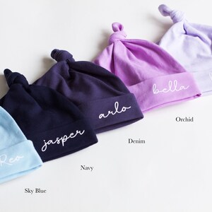 May include: Five personalized baby hats in different colors: sky blue, navy, denim, orchid, and lavender. The hats are made of soft cotton and have a knotted top. Each hat is embroidered with a different name: Rea, Jasper, Arlo, Bella, and Harper.