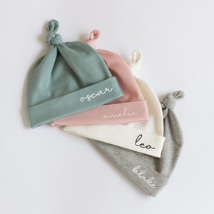 May include: Four personalised baby hats in different colours: teal, pink, white and grey. The hats are knotted at the top and have names embroidered on them: Oscar, Amelia, Leo and Blake.