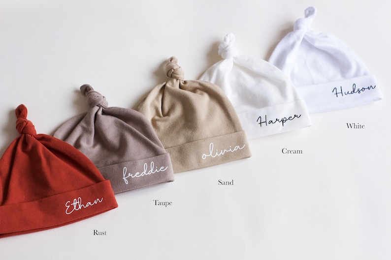 May include: Five different colored knotted baby hats with personalized names embroidered on them. The colors are rust, taupe, sand, cream, and white. The names are Ethan, Freddie, Olivia, Harper, and Hudson.