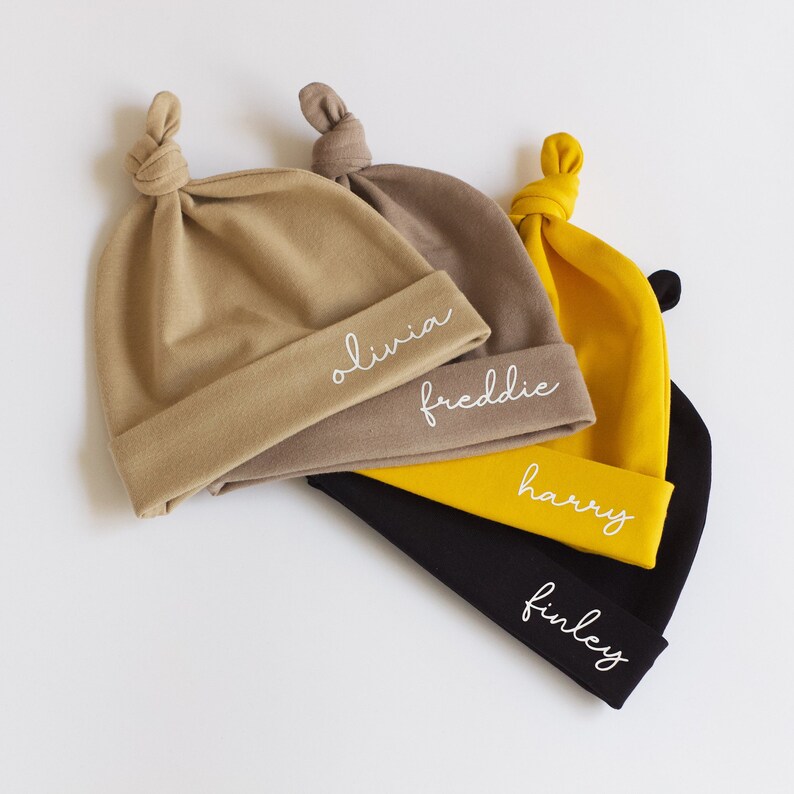 Peut inclure: Quatre bonnets pour b&eacute;b&eacute; nou&eacute;s en beige, jaune et noir avec des noms brod&eacute;s en blanc&nbsp;: Olivia, Freddie, Harry et Finley.