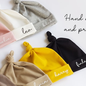 Peut inclure: Une collection de dix bonnets pour b&eacute;b&eacute;s faits &agrave; la main et imprim&eacute;s, dans diff&eacute;rentes couleurs, chacun avec un nom diff&eacute;rent brod&eacute; sur le bord. Les bonnets sont faits d'un tissu doux et extensible et ont un n&oelig;ud sur le dessus.