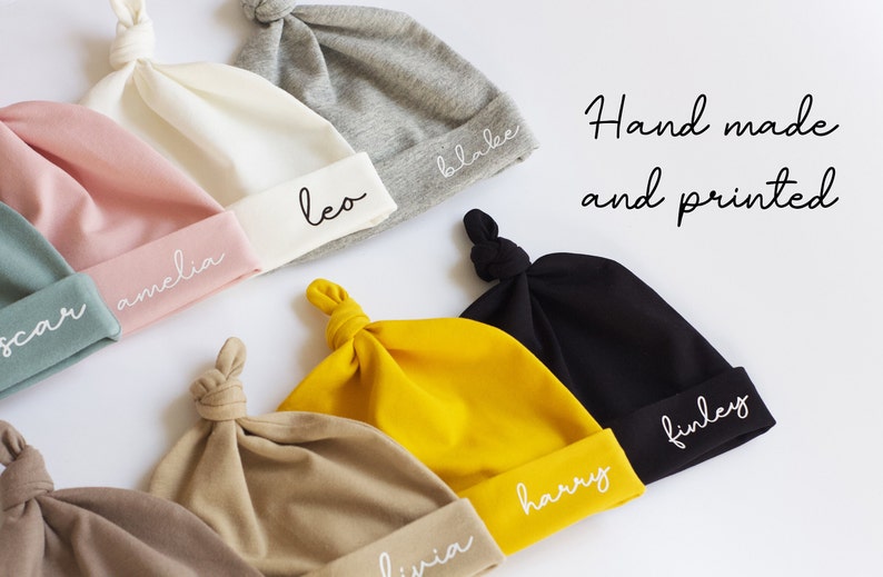 Peut inclure: Une collection de dix bonnets pour b&eacute;b&eacute;s faits &agrave; la main et imprim&eacute;s, dans diff&eacute;rentes couleurs, chacun avec un nom diff&eacute;rent brod&eacute; sur le bord. Les bonnets sont faits d'un tissu doux et extensible et ont un n&oelig;ud sur le dessus.