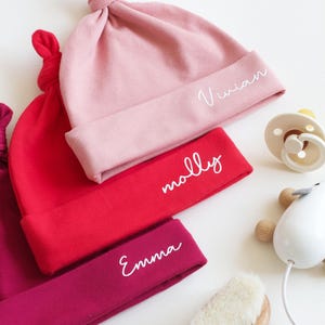 May include: Three baby hats in pink, red, and burgundy with white embroidered names: Vivian, Molly, and Emma.