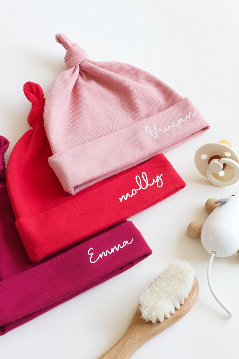 May include: Three baby hats in pink, red, and burgundy with white embroidered names: Vivian, Molly, and Emma.