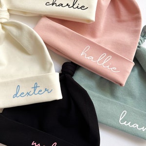 May include: Five different colored baby hats with knotted tops. The hats are white, pink, black, and light blue. Each hat has a name embroidered on it: Charlie, Hallie, Dexter, Wednesday, and Luan.