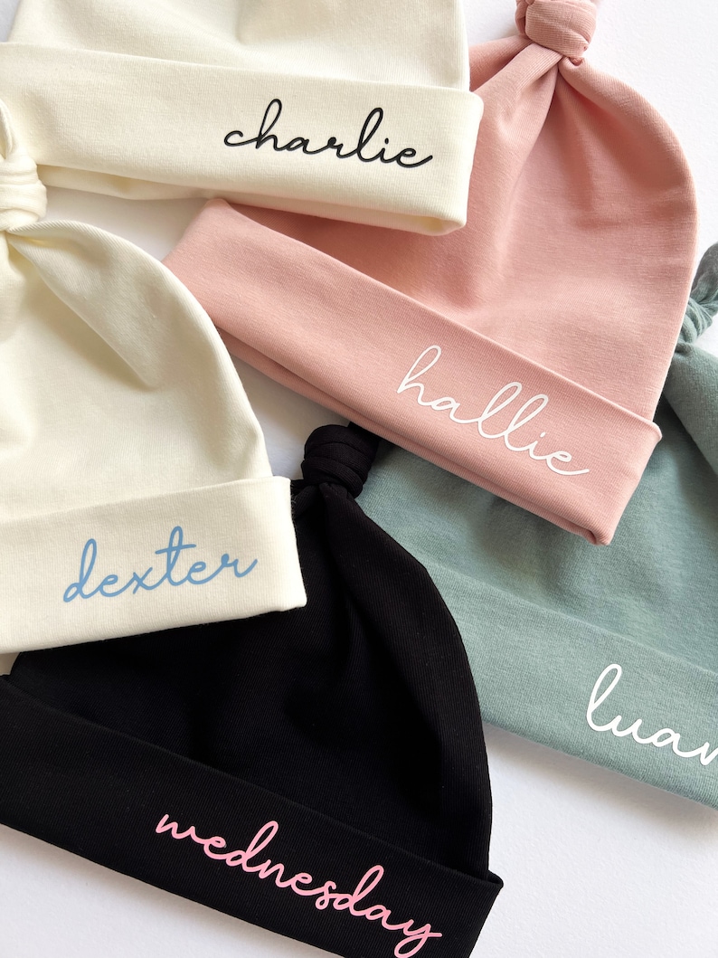 May include: Five different colored baby hats with knotted tops. The hats are white, pink, black, and light blue. Each hat has a name embroidered on it: Charlie, Hallie, Dexter, Wednesday, and Luan.