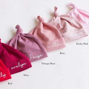 May include: Six different colored baby hats with knotted tops. The hats are labeled with the names "Molly", "Evelyn", "Paige", "Willow", "Robyn", and "Candyfloss". The colors of the hats are red, wine, vintage rose, rose, dusky pink, and candyfloss.