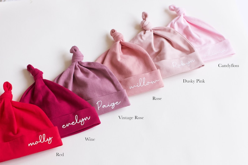 May include: Six different colored baby hats with knotted tops. The hats are labeled with the names "Molly", "Evelyn", "Paige", "Willow", "Robyn", and "Candyfloss". The colors of the hats are red, wine, vintage rose, rose, dusky pink, and candyfloss.