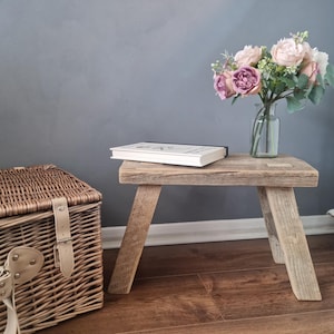 May include: A rustic wooden stool with a natural wood finish. The stool has a simple design with four legs and a rectangular top. A book is placed on top of the stool. A bouquet of pink flowers in a clear glass vase is on the right side of the stool.
