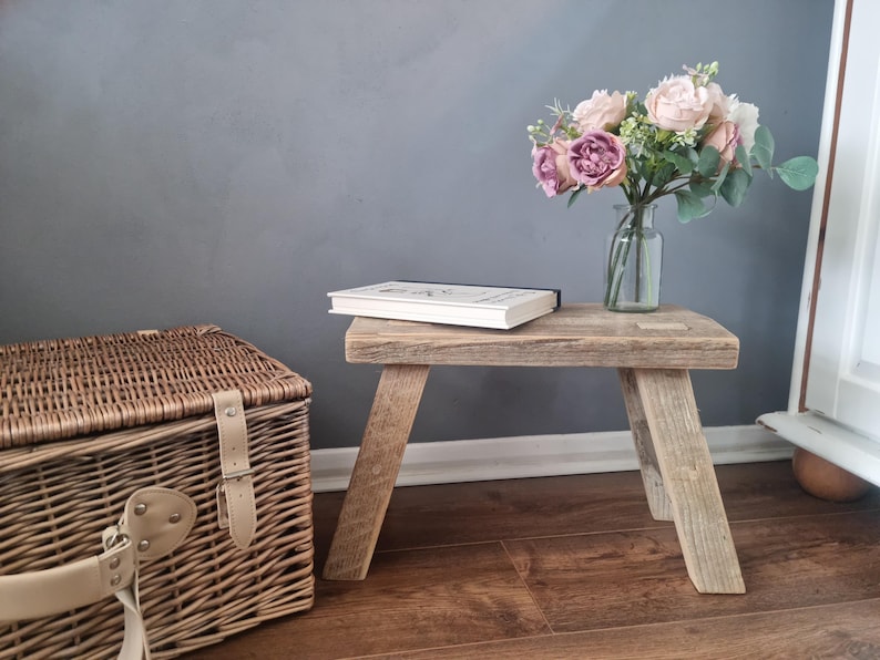 May include: A rustic wooden stool with a natural wood finish. The stool has a simple design with four legs and a rectangular top. A book is placed on top of the stool. A bouquet of pink flowers in a clear glass vase is on the right side of the stool.