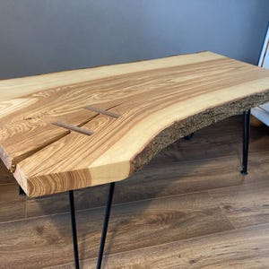 May include: A natural wood coffee table with a live edge top and black hairpin legs. The wood has a unique grain pattern and is finished in a natural color.