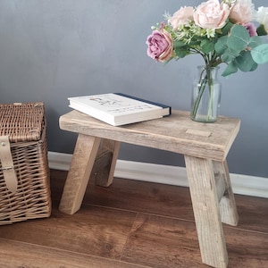 May include: A rustic wooden stool with a natural wood finish. The stool has a book on top and a vase of pink flowers next to it. The stool is sitting on a wooden floor.