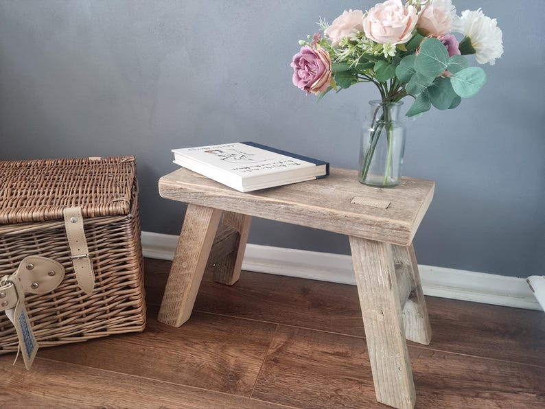 May include: A rustic wooden stool with a natural wood finish. The stool has a book on top and a vase of pink flowers next to it. The stool is sitting on a wooden floor.