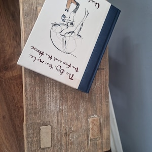 May include: A white book with a black and white illustration of a person riding a horse. The book's title is "The Boy, the Fox and the Horse" by Charlie Mackesy. The book is sitting on a wooden surface.