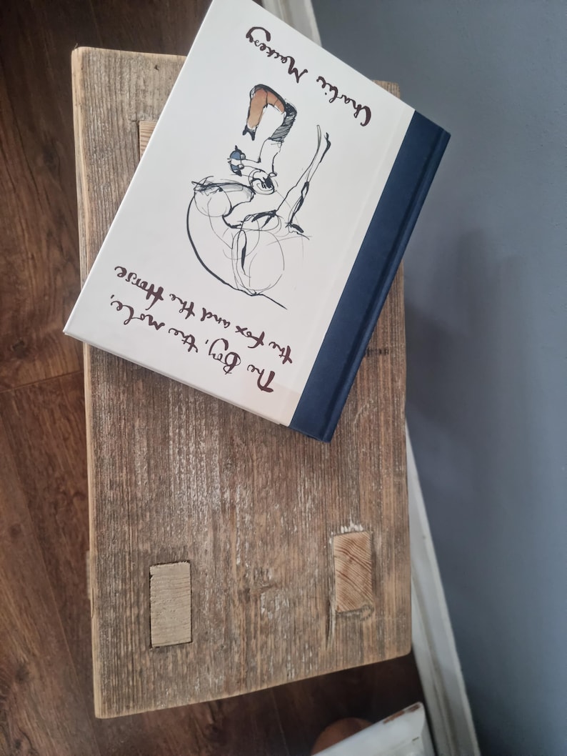 May include: A white book with a black and white illustration of a person riding a horse. The book's title is "The Boy, the Fox and the Horse" by Charlie Mackesy. The book is sitting on a wooden surface.