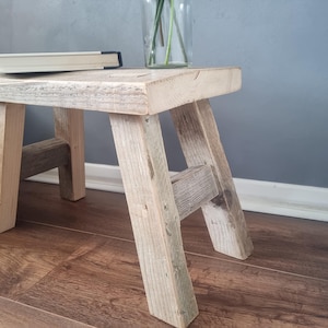 May include: A rustic wooden stool with a natural wood finish. The stool has a simple design with four legs and a rectangular top. The stool is made from reclaimed wood and has a weathered look.