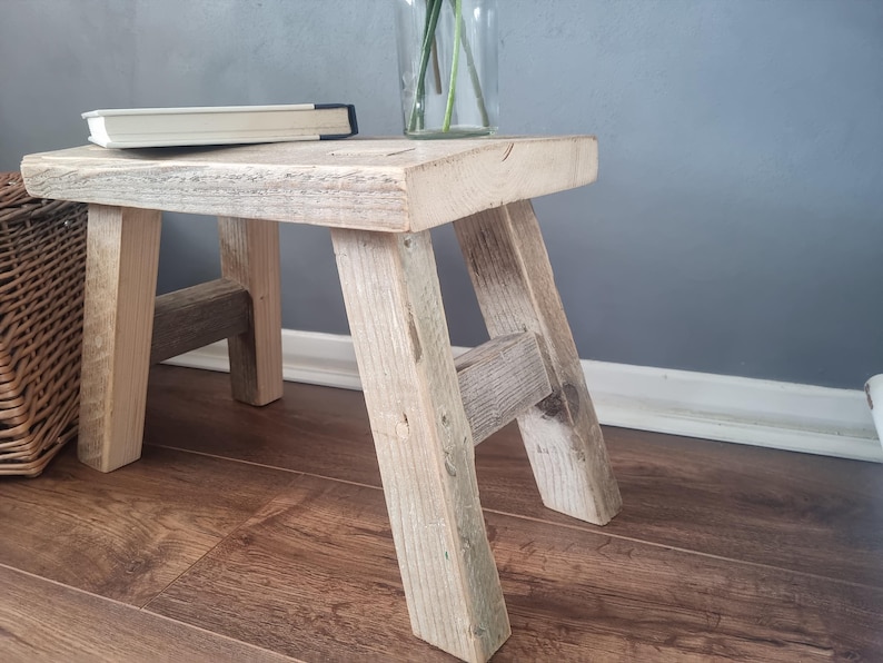 May include: A rustic wooden stool with a natural wood finish. The stool has a simple design with four legs and a rectangular top. The stool is made from reclaimed wood and has a weathered look.