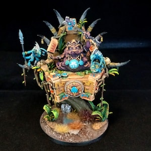 May include: A detailed miniature figurine of a fantasy throne. The throne is made of tan stone and adorned with blue and turquoise accents. A large purple creature sits on the throne, surrounded by smaller blue figures and lush green foliage. The base is brown.