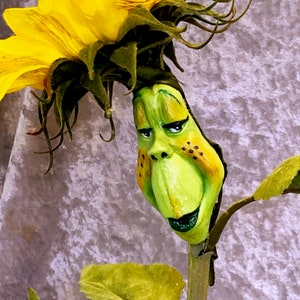 May include: A handcrafted sunflower sculpture with a whimsical design. The bright yellow petals surround a green, expressive face with a downturned mouth and freckles. The stem and leaves are green, set against a textured, lavender background.