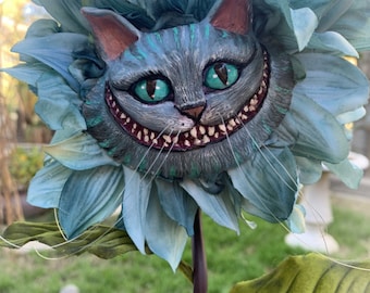 May include: A blue flower with a Cheshire Cat face in the center. The cat has a wide grin with sharp teeth and green eyes. The flower is made of fabric and has a long stem.