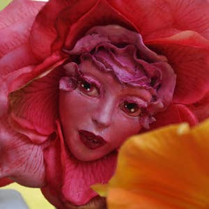 May include: A pink and red fabric flower with a human face in the center. The face has pink skin, brown eyes, and red lips. The flower is surrounded by a yellow flower.