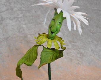May include: A whimsical green and white flower with a smiling face, crafted from fabric and displayed on a green base with moss.