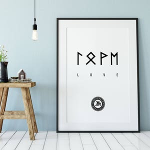 Puede incluir: Una impresión enmarcada en blanco con texto negro y un marco negro. La impresión presenta símbolos rúnicos sobre la palabra "LOVE". Un pequeño taburete de madera sostiene una jarra negra con flores, una casa en miniatura y velas.
