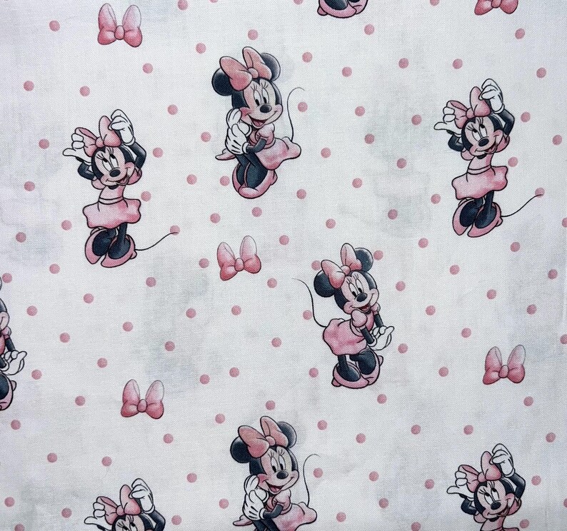 K&ouml;nnte beinhalten: Wei&szlig;er Stoff mit einem sich wiederholenden Muster von Minnie Mouse in rosa Kleidern und Schleifen sowie rosa Tupfen. Die Zeichentrickfigur ist in verschiedenen Posen zu sehen, mit schwarzen Ohren und einer rosa Schleife.
