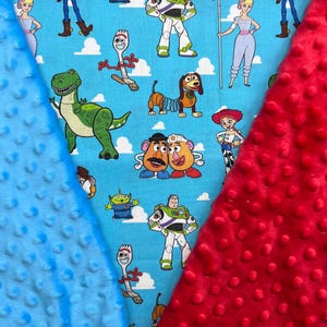 Op de afbeelding: Een babydeken met een lichtblauwe achtergrond en stripfiguren. De deken heeft een rode minky dot stof aan de rechterkant en een blauwe minky dot stof aan de linkerkant. De stripfiguren zijn onder andere Buzz Lightyear, Woody en Rex.