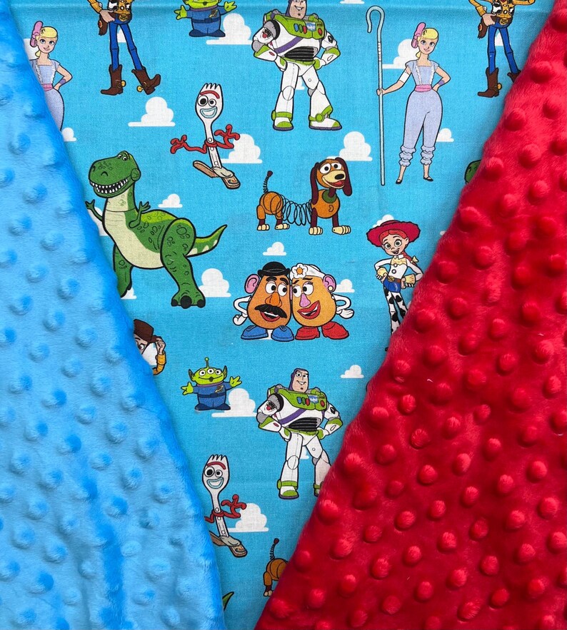 Op de afbeelding: Een babydeken met een lichtblauwe achtergrond en stripfiguren. De deken heeft een rode minky dot stof aan de rechterkant en een blauwe minky dot stof aan de linkerkant. De stripfiguren zijn onder andere Buzz Lightyear, Woody en Rex.