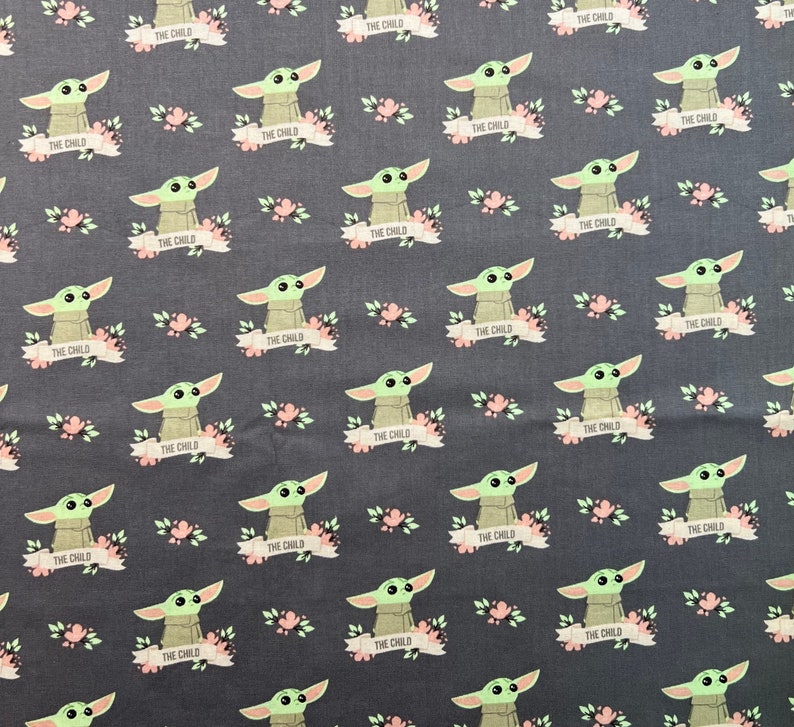May include: A dark gray fabric with a repeating pattern of a green alien character with big eyes and the text "THE CHILD" on a white banner. The alien is surrounded by pink flowers.