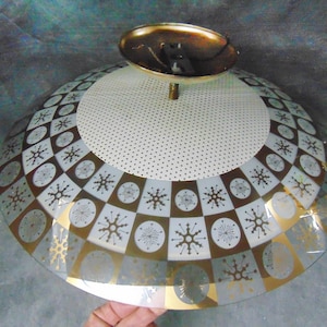May include: Vintage mid-century modern ceiling light fixture. The glass shade features a geometric pattern of squares, circles, and snowflake-like designs in gold, white, and brown. The fixture has a brass-colored top.