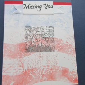 Op de afbeelding: Een wenskaart met een verweerde vlagpatroon in rood, wit en blauw. Een centraal vierkant blok toont een zwarte inktillustratie van een adelaarskop omringd door woorden als loyaliteit, moed en eer. De tekst Missing You staat op een witte banner aan de bovenzijde.