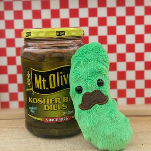 May include: A jar of Mt. Olive Kosher Baby Dills pickles next to a plush green pickle toy with a brown mustache and black eyes. The jar is labeled with text and a yellow lid. The background is a red and white checkered pattern.