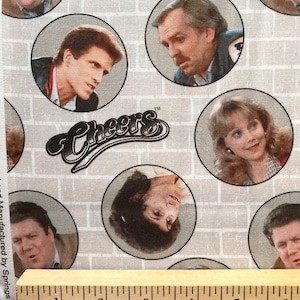 May include: A white fabric with a repeating pattern of gray brick and circles. Each circle contains a different character from the TV show "Cheers" with the show's logo in the center. The logo is black and white and says "Cheers" with a trademark symbol.